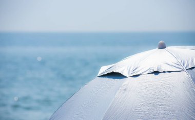 Ön planda soluk pembe çizgili plaj şemsiyesi, mavi deniz arka planına karşı..