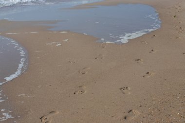 Kumsalda ayak izleri. Kumdaki ayak izleri bir deniz dalgasına karşı. Güneşli bir günde altın kum, sahil, dalga ve ayak izleri..