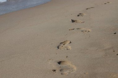 Kumsalda ayak izleri. Kumdaki ayak izleri bir deniz dalgasına karşı. Güneşli bir günde altın kum, sahil, dalga ve ayak izleri..