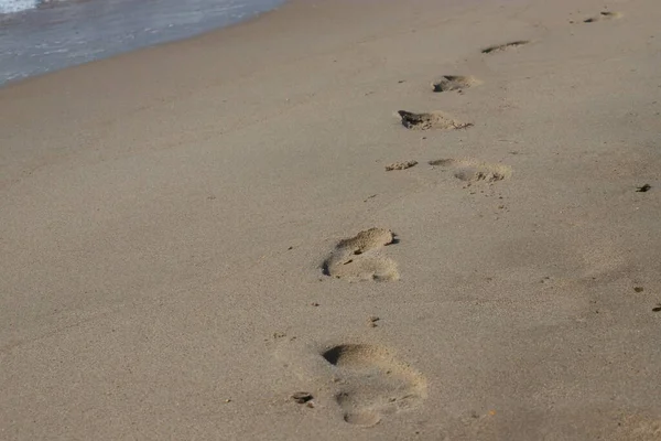 Kumsalda ayak izleri. Kumdaki ayak izleri bir deniz dalgasına karşı. Güneşli bir günde altın kum, sahil, dalga ve ayak izleri..