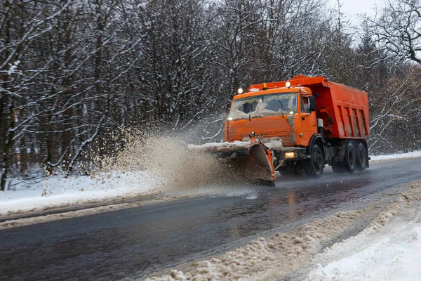 A large car with a plow clears the road from snow. Orange cargo special equipment is struggling with the elements in winter. Removing the effects of the snowstorm. Difficulties in traffic.Frozen water.