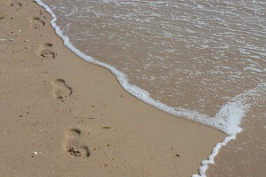 Kumsalda ayak izleri. Kumdaki ayak izleri bir deniz dalgasına karşı. Güneşli bir günde altın kum, sahil, dalga ve ayak izleri..