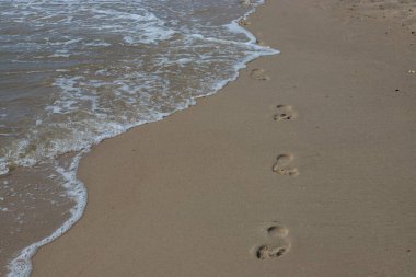 Kumsalda ayak izleri. Kumdaki ayak izleri bir deniz dalgasına karşı. Güneşli bir günde altın kum, sahil, dalga ve ayak izleri..