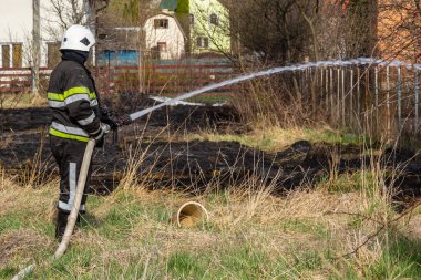 spring fire, burning dry grass near buildings in the countryside. Firefighter extinguishes the flame. Environmental disaster.