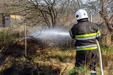 spring fire, burning dry grass near buildings in the countryside. Firefighter extinguishes the flame. Environmental disaster.