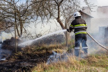 spring fire, burning dry grass near buildings in the countryside. Firefighter extinguishes the flame. Environmental disaster.