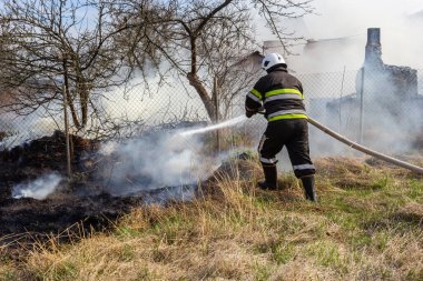 spring fire, burning dry grass near buildings in the countryside. Firefighter extinguishes the flame. Environmental disaster.