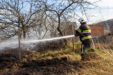 spring fire, burning dry grass near buildings in the countryside. Firefighter extinguishes the flame. Environmental disaster.
