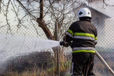 spring fire, burning dry grass near buildings in the countryside. Firefighter extinguishes the flame. Environmental disaster.