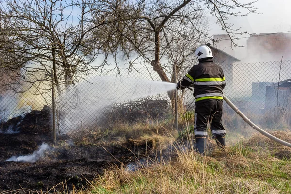 spring fire, burning dry grass near buildings in the countryside. Firefighter extinguishes the flame. Environmental disaster.