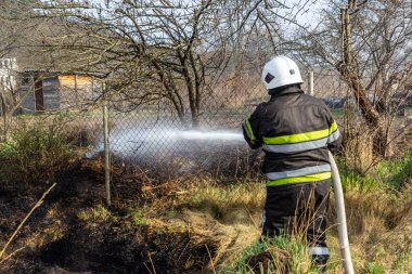 spring fire, burning dry grass near buildings in the countryside. Firefighter extinguishes the flame. Environmental disaster.