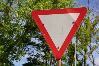Yol tabelasına yakın çekim. Kırmızı ve beyaz üçgen. Arkaplanda yeşil ağaçlar ve mavi gökyüzü.