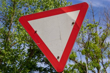 Yol tabelasına yakın çekim. Kırmızı ve beyaz üçgen. Arkaplanda yeşil ağaçlar ve mavi gökyüzü.