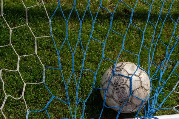 old soccer ball in the net on the background of grass soccer field. Summer sunny day.