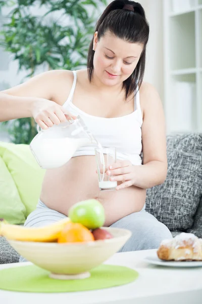 怀孕的女人坐在沙发上，把牛奶倒入一杯 — 图库照片