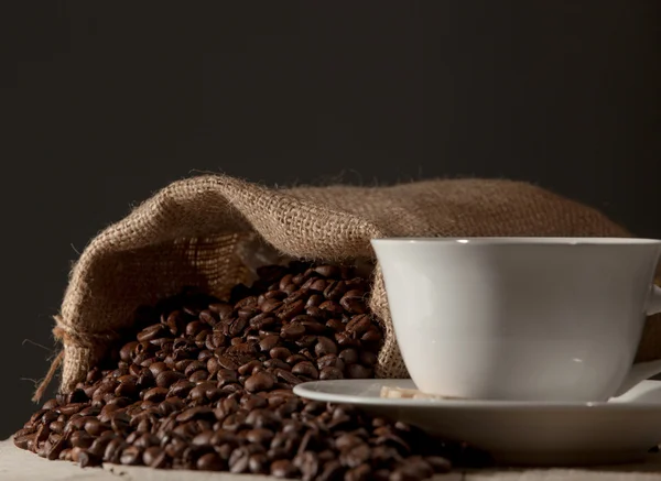Koffiekopje met jute zak van geroosterde bonen tegen donkere hout b — Stockfoto