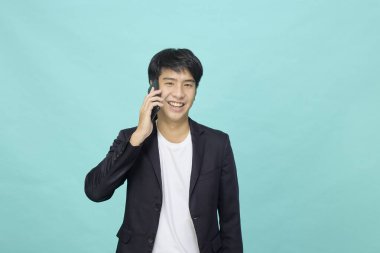 Genç, gülümseyen, takım elbiseli yakışıklı Asyalı adam açık mavi izole stüdyo arka planında telefonda konuşuyor.
