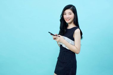 Genç Asyalı bayan mavi arkaplandaki kameraya bakarak pozitif bir ifadeyle cep telefonu kullanıyor. Mutlu, mutlu, mutlu kadın başarıya sevinir..