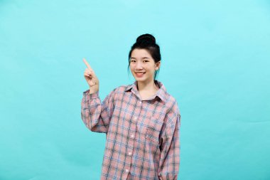 Genç Asyalı bir kadın ekose gömlek giymiş reklamlara bakıyor ve mavi arka planda mutlu ve kendinden emin hissediyor.