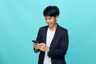 Yarı resmi takım elbiseli gülümseyen yakışıklı Asyalı bir adamın portresi açık mavi izole stüdyo arka planında cep telefonu kullanıyor.