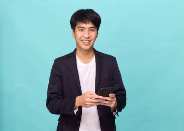 Yarı resmi takım elbiseli gülümseyen yakışıklı Asyalı bir adamın portresi açık mavi izole stüdyo arka planında cep telefonu kullanıyor.