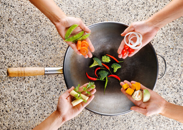 Four hands puts fresh vegetables in the wok. Cooking concept