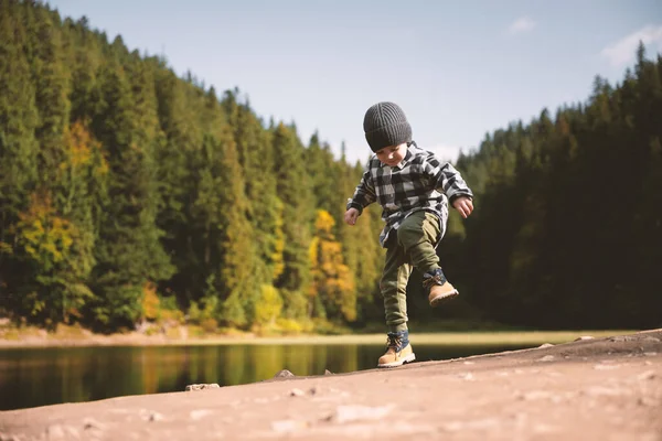 Criança pequena em uma camisa xadrez e chapéu cinza na costa do lago da floresta — Fotografia de Stock