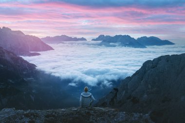 Dolomites dağlarındaki uçurumun kenarında sislerin üzerinde oturan bir turist.