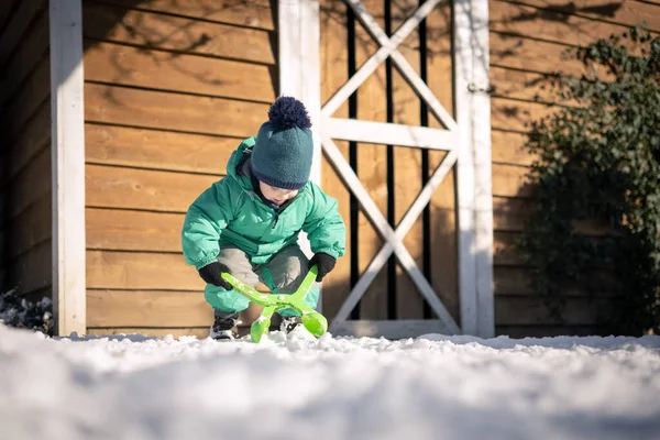 Kleines Kind in grüner Jacke und Strickmütze bastelt Schneebälle in der Nähe des Hauses — Stockfoto