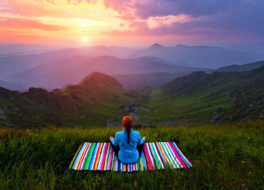 Yogacı kız yüksek dağlardaki çimlerde meditasyonda çizgili minderde oturuyor ve gökyüzü ışınlar ve bulutlarla kaplı. Bahar sabahında gün doğumu. Duvar kağıdı arka planı. Doğal manzara..