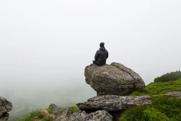 身穿黑色夹克的矮胖男子正坐在悬崖边上 多雾天气下的山区景观 — 图库照片