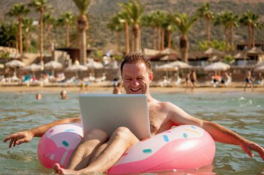 Her yerden çalışma ve uzaktan işgal konsepti. Tropikal denizlerdeki şişme halka üzerine dizüstü bilgisayarla çalışan çok mutlu bir iş adamı.. 