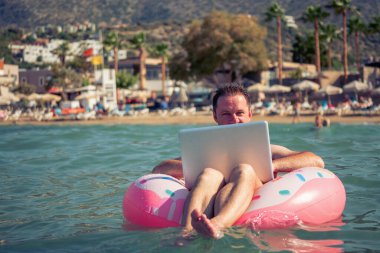 Her yerden ve her zaman çalışmak, uzak işgal konsepti. Tropik denizdeki şişme halka üzerinde dizüstü bilgisayarla çalışan iş adamının portresi. 