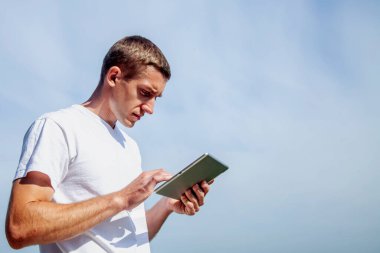 Her yerde ve her zaman çalışıyorum. Tablet kullanan ve açık havada mavi gökyüzünün arka planına karşı çalışan yakışıklı bir iş adamı. Tasarım için alanı kopyala.