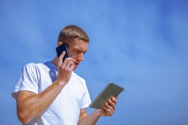 Tablet kullanan yakışıklı bir iş adamı ve telefonda mavi gökyüzünün arka planına karşı konuşuyor. Tasarım için alanı kopyala.