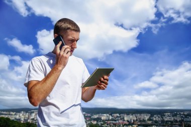 Tablet kullanan genç ve yakışıklı işadamının portresi ve telefonda şehrin panoramik manzarasına karşı konuşmak. Metin iletisi için ayarlayın. 