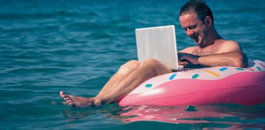 Dizüstü bilgisayarla denizde şişme halka üzerinde çalışan serbest çalışan bir adam. Seçici odaklanma. Tasarım için alanı kopyala.