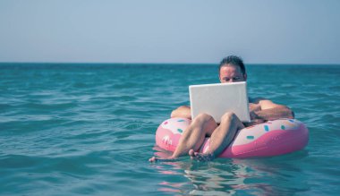 Her yerde ve her zaman çalışmak. Genç adam dizüstü bilgisayarda deniz suyunda şişme halkayla çalışıyor. Boşluğu kopyala