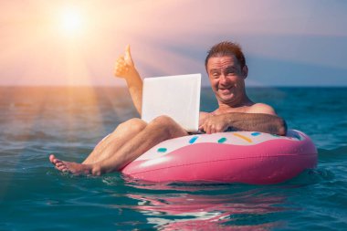 Her yerde ve her zaman çalışmak. Mutlu adam serbest iş adamı laptopun başında denizin sularında şişme halkayla çalışıyor.. 