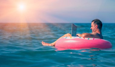 Her yerde ve her zaman çalışmak. Genç adam dizüstü bilgisayarda deniz suyunda şişme halkayla çalışıyor. Boşluğu kopyala