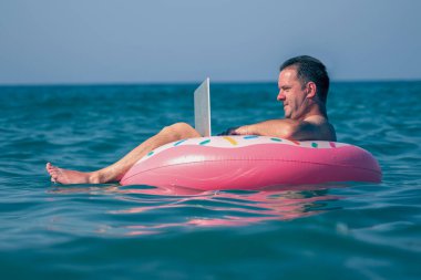 Her yerde ve her zaman çalışmak. Genç adam dizüstü bilgisayarda deniz suyunda şişme halkayla çalışıyor. Yatay resim.