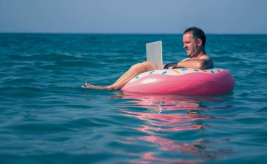 Her yerde ve her zaman çalışmak. Genç adam dizüstü bilgisayar başında denizin sularında şişme halka üzerinde çalışıyor.