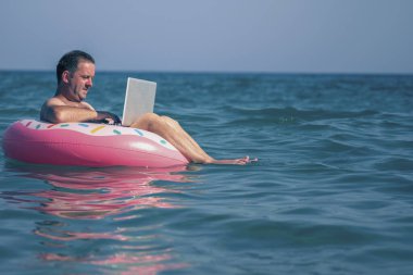 Erkek gezgin deniz ve okyanus sularındaki şişme halkalar üzerinde dizüstü bilgisayar kullanıyor ve çalışıyor. Boşluğu kopyala.