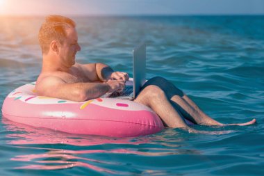 Genç adam deniz suyunda şişirilebilir halka üzerine bir dizüstü bilgisayarla tatilde çalışıyor.. 