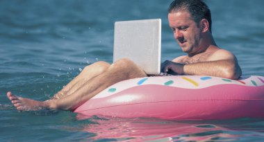 Tatilde deniz ve okyanus sularında şişme halka üzerinde dizüstü bilgisayarla çalışan erkek girişimci portresi. Yatay resim.