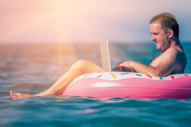 Erkek girişimci, deniz ve okyanus sularında şişme halka üzerine dizüstü bilgisayarla çalışıyor.