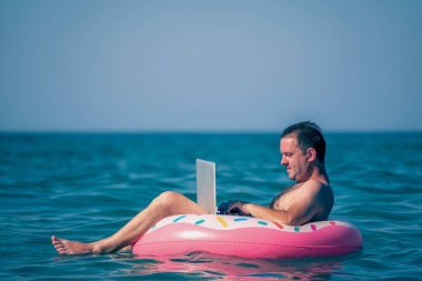 Erkek girişimcinin portresi deniz ve okyanus sularında şişirilebilir halka üzerinde dizüstü bilgisayarla çalışıyor. Boşluğu kopyala.