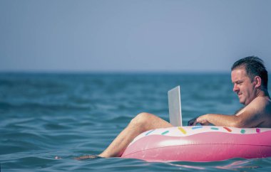 Her yerde ve her zaman çalışmak. Sudaki şişme halka üzerinde dizüstü bilgisayar kullanan genç bir adam çalışıyor. Serbest çalışma, tatiller, mesai, sosyal mesafeler. Metin için alanı kopyala