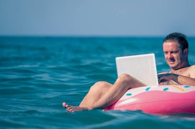 Her yerde ve her zaman çalışmak. Sudaki şişme halka üzerinde dizüstü bilgisayar kullanan genç bir adam çalışıyor. Serbest çalışma, tatiller, mesai, sosyal mesafeler. Uzayı kopyala Yatay resim.