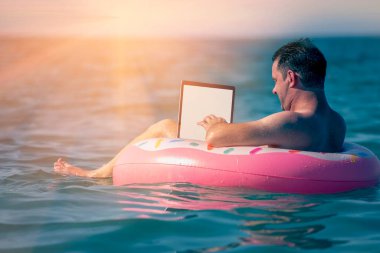 Her yerde ve her zaman çalışmak. Sudaki şişme halka üzerinde dizüstü bilgisayar kullanan genç bir adam çalışıyor. Serbest çalışma, tatiller, mesai, sosyal mesafeler. Yatay resim.